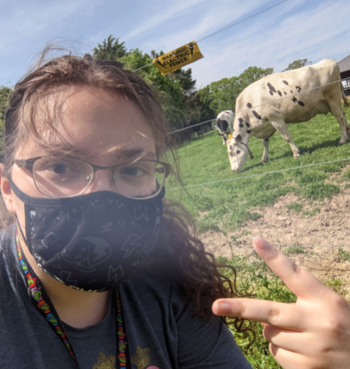 Blade standing in front of a wire fence. She has her brown hair tired back in a ponytail with a black facemask on. She is giving a peace sign. Behind her is a white cow with black spots.